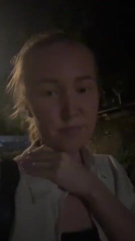 A woman with blonde hair pulled back is standing outside at night. She is wearing a light-colored button-up shirt over a dark tank top. She is holding her right hand to her neck and throat area. The background is dark with some blurry lights and vegetation. The woman is looking towards the camera.