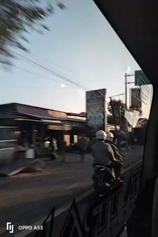 A video captures a street scene from a moving vehicle. The view is primarily through a window, with part of the vehicle's interior visible at the bottom and right edges. The street appears to be a rural or semi-urban area, with buildings lining one side and a dirt or unpaved road. Overhead power lines are visible. The sky is a light blue, suggesting it might be early morning or late afternoon.

In the foreground, two people are riding a motorcycle, traveling away from the camera. The person in front is wearing a white helmet, and the person behind is wearing a dark helmet.

Further down the street, a large, dark-colored truck with a tall, open-top bed is moving towards the camera, its bed appearing to be empty.

Several buildings are visible along the street. Some appear to be shops or small businesses with closed shutters. There are also some billboards or large signs erected along the side of the road. One large billboard has a picture of people in white garments. Another, partially obscured, seems to have a dark design.

As the vehicle continues, a dark-colored car is parked on the side of the road, and two individuals are near it. Another motorcycle is visible further down the street. A satellite dish is mounted on top of one of the buildings. A tall, slender structure, possibly a telecommunications tower, is visible in the distance.

The video captures the movement of vehicles and people along the street, creating a sense of everyday life in this location. The lighting suggests a time of day when the sun is low.