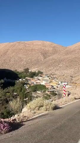 This is a wide shot of a small village nestled in a valley between two arid, rocky hills. The sky is a clear, deep blue. A paved road curves into the foreground from the right. In the middle ground, a cluster of small, colorful buildings makes up the village. Some of the buildings are single-story, while others have multiple stories. There are trees and vegetation around the village, especially on the left side of the valley. A red and white striped pole, likely a traffic marker, stands on the right side of the road. The overall impression is of a remote, sun-drenched settlement in a mountainous desert landscape.