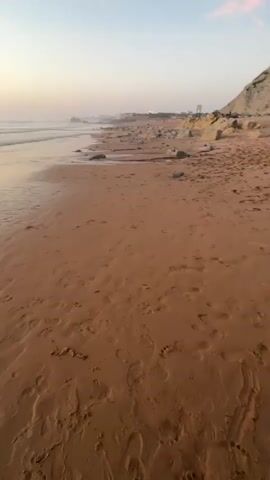 The video shows a beach at sunset. The sand is wet and has footprints on it. The ocean has small waves. The sky is a mix of blue, pink, and orange, with some clouds. The sun is low on the horizon, casting a warm glow over the scene. There are some rocks and cliffs in the distance. The video is shot from a low angle, close to the ground.