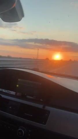 The video shows a view from the inside of a car looking out the windshield at a sunset. The sun is low on the horizon, casting a warm orange glow across the sky and clouds. The car appears to be on a highway, with other vehicles visible in the distance. The dashboard of the car is visible in the foreground.