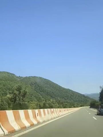 This is a first-person perspective video taken from a moving vehicle, likely a car, driving on a paved road. The road is flanked by a concrete barrier with alternating orange and white sections. On the left side of the road, there are lush green hills covered in trees, with a clear blue sky above. On the right side of the road, a dark-colored car with a roof rack is visible in the distance. The video appears to be shot during daylight, with bright sunlight illuminating the scene. The camera is pointed slightly forward and to the right, capturing the road ahead and the surrounding landscape. There are no discernible people visible in the frame. The overall impression is one of travel through a scenic, mountainous region.