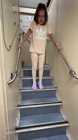 A young girl with long brown hair is descending a staircase. She is wearing a light-colored t-shirt with a pink and orange Adidas logo and matching leggings with a patterned design. She has on purple clogs. The staircase has blue carpeted steps with a dark strip on each edge and metal handrails on both sides. The walls of the stairwell are a light beige color. The girl is holding onto the handrails with both hands as she walks down the stairs. She appears to be looking down and has a surprised or playful expression on her face.