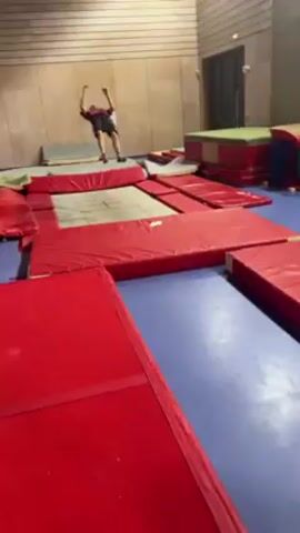 A person practices gymnastics indoors on a trampoline surrounded by red mats. The individual performs a backflip with a twist, landing successfully. The room has blue flooring, paneled walls, and gymnastic equipment along the sides. The lighting is bright.