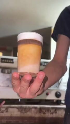 A close-up shot shows a hand holding a layered coffee drink in a clear glass. The drink has distinct layers of white foam on top, followed by a dark brown layer, a lighter brown layer, and a creamy white layer at the bottom. In the background, a person wearing a dark blue shirt and jeans is partially visible. Behind them, a white espresso machine is visible. The environment appears to be indoors, possibly a cafe or a kitchen, with warm lighting.