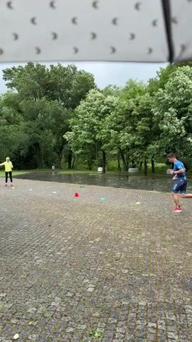 A man in athletic wear sprints through a park in the rain, weaving around small cones. He is wearing a blue shirt, dark shorts, and red and blue socks. A woman in a yellow jacket and red pants is positioned further down the path, possibly giving instructions. Another person wearing a pink raincoat stands in the foreground, observing the man's run. The ground is paved with stone blocks, and the park is green with trees. The camera is looking out from under the cover of an umbrella.