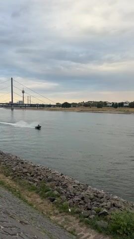 A jet ski speeds across a wide river, leaving a white wake. In the background, a suspension bridge spans the river, and beyond it, a city skyline with a prominent tower is visible. The sky is overcast with clouds. The riverbank in the foreground is lined with rocks and vegetation.