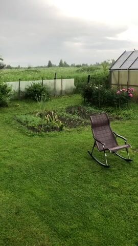 A wide shot captures a backyard scene under an overcast sky, with rain visible as streaks against the cloudy background. In the foreground, a well-maintained green lawn stretches out, interrupted by a brown rocking chair positioned on the grass. To the left, a garden bed contains various plants, including some tall, green stalks and smaller leafy greens, with a few visible flower stalks. A white plastic fence borders the garden area and extends further back. Beyond the garden, a greenhouse with a translucent roof and walls stands to the right. In the background, a vast field of tall green grass and some bushes can be seen, extending towards a distant horizon under the grey sky. The overall impression is that of a quiet, rainy day in a rural or suburban garden.