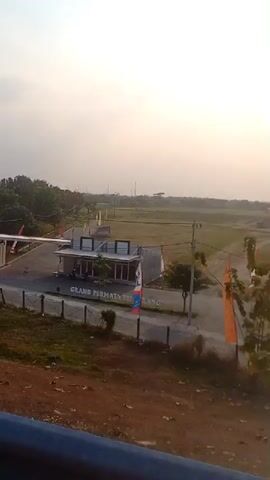The video shows a slightly hazy view of a residential complex entrance, possibly Grand Permata Telang. The entrance features a modern building with a white awning and a sign that reads "GRAND PERMATA TELANG". There are several flags and decorative elements around the entrance. A dark-colored car is visible driving away from the entrance. The surrounding area is mostly green with trees and open fields, suggesting a rural or suburban setting. The sky is overcast with the sun low on the horizon, indicating either early morning or late afternoon. The foreground is a bit blurry, showing a dirt embankment and a dark railing. The overall impression is of a developing housing project.