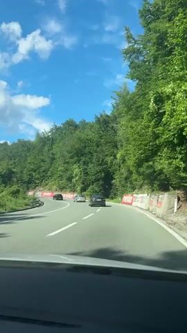 The video shows a car's point-of-view driving on a paved road through a forested area. Several cars are visible ahead on the road, which curves to the right. The road has white lane markings. On the right side of the road, there are red and white barriers with some graffiti on them. The sky is blue with white clouds. The trees are dense and green, lining both sides of the road.