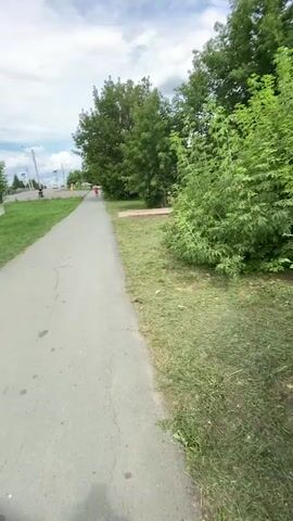 A first-person perspective video begins with a sidewalk and grass, with trees and a building in the background. A person runs in the distance. The camera pans to the right, showing more grass and trees. It then pans left and shows a person on a bicycle in the distance. The camera continues to pan left, and the face of the person recording the video is visible, wearing a white hat and a white top. The video ends with a shot of the person recording, with trees and a building in the background.