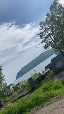 The video begins with a shot looking across a body of water towards a forested hill. The sky is blue with scattered clouds.  As the video progresses, the camera pans to the right, revealing trees and a small building. The scene then shifts to a view from inside a vehicle, looking out the windshield. The vehicle is traveling on a paved road with a white line marking the edge. Trees and a guardrail are visible along the side of the road. Other vehicles, including a dark SUV and a smaller car, are ahead on the road. The sky remains blue with white clouds.