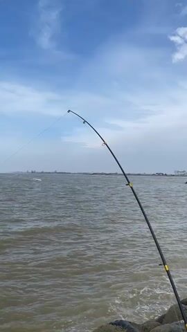 A fishing rod is held at an angle over the water. The rod is black with yellow accents. A thin fishing line extends from the tip of the rod into the water. The water is choppy and a distant shoreline is visible under a cloudy blue sky. The camera appears to be positioned behind the fishing rod, looking out over the water.