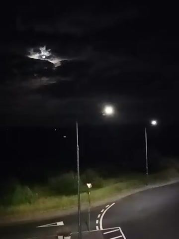 The video shows a nighttime outdoor scene. Two streetlights illuminate a dark asphalt road with white markings. In the background, there is dark vegetation and a faint horizon line. The sky is dark with clouds. The moon is visible through a gap in the clouds, casting a faint light. There are no people or animals visible in the video.
