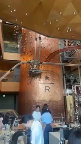 The camera pans upwards inside what appears to be a Starbucks Reserve store. A large, cylindrical, copper-colored structure dominates the scene, adorned with a Starbucks logo and the letter "R". Tubes and mechanical elements extend from it. Below, baristas work behind a counter. The ceiling is a geometric pattern with recessed lighting. People are standing around and interacting with the baristas.