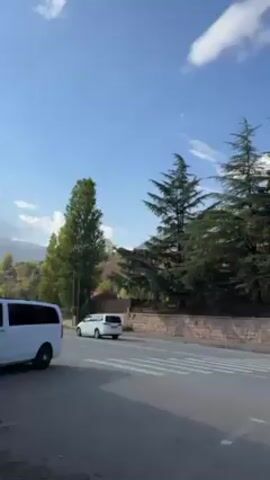 The camera pans from left to right, showing a roadside scene under a bright blue sky with scattered clouds. On the left, a white van is parked and a white car drives past. A wide road with a crosswalk is visible, and a gray car travels down it. Beyond the road, a low stone wall separates it from a wooded area filled with tall green trees and a few trees with yellowish leaves. In the background, a mountain is partially visible. Power lines and a streetlight run alongside the road. A pedestrian walks along the sidewalk to the right.