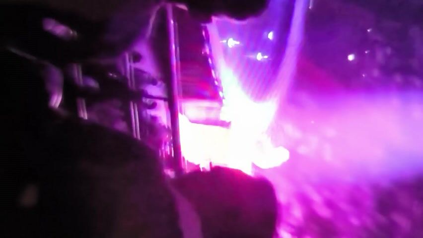 The video shows a person in mid-air, appearing to be suspended by wires as part of a performance or stunt. The subject is wearing a light-colored, possibly white or silver, outfit with dark accents. Their arms are extended outwards, and their body is positioned horizontally. The background is a mix of deep purple and bright pink, suggesting stage lighting or a similar artificial environment.

A prominent object in the scene is a large, vertical, red structure with a white element on its surface, which the performer seems to be interacting with or flying past. Below the performer, a large, arched structure, illuminated with white and purple light, is visible. This archway appears to be made of a material with a textured or woven surface, possibly fabric or netting, stretched over a frame. Another figure, wearing darker clothing, is partially visible near the base of the red structure, possibly part of the technical crew or another performer.

The overall impression is of a dramatic and dynamic performance, likely taking place indoors on a stage or at an event. The lighting creates a vibrant and possibly theatrical atmosphere. There is no visible text in the video. The time of day cannot be determined due to the artificial lighting. No weather or specific location cues beyond the provided context of Oktyabrsky, Russia, are discernible from the visual information. The performer's pose and the context suggest an action sequence, possibly involving acrobatics or a visual effect.