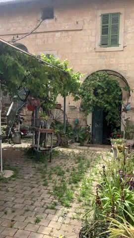 The camera slowly pans across a charming, overgrown courtyard. The ground is paved with brick, and scattered patches of grass and weeds grow between the stones.  A weathered stone building forms the backdrop, with a doorway framed by climbing vines.  A structure covered in grape vines provides shade over a dining area, which has a wooden table and chairs.  The yard is filled with an assortment of plants in pots, including cacti, and other greenery.  Various objects are scattered around, including a barbecue grill, a metal milk jug, and garden tools. The overall atmosphere is rustic and inviting.