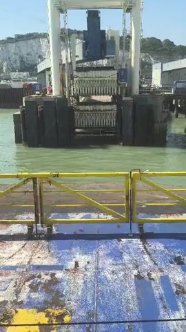 A video shows a ferry approaching a dock. The ferry has a blue and yellow deck. A yellow gate is in the foreground. In the background, a large mechanical structure with a series of poles is visible. The structure is partially submerged in green water. White cliffs and buildings are visible in the distance. The sky is blue and clear.