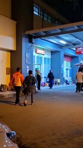 At night, a group of people are standing outside what appears to be an emergency room entrance of a hospital. An ambulance is parked with its rear doors open, and two individuals are seen near it. One person is wearing a white shirt and is facing away from the camera, standing beside the open ambulance. Another person in a dark outfit is walking towards the ambulance. A third person in an orange jacket is walking away from the ambulance towards the left of the frame. Several other individuals are seated or standing near the entrance, some with bags beside them. A sign above the entrance reads "EMERGENCY ONLY". The overall scene suggests a busy or possibly chaotic situation at the hospital entrance, with people waiting or tending to something related to the ambulance.