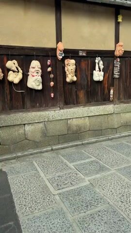 The video shows a street scene in Japan. The first shot is of a traditional wooden building with several decorative masks hanging on its exterior. The camera then pans to two women walking away from the camera, dressed in traditional Japanese kimonos. The woman on the left is wearing a light pink kimono with a ruffled obi, and the woman on the right is wearing a dark blue kimono with a floral pattern and a light-colored obi, carrying a white handbag. In the background, other people are visible, and a multi-tiered pagoda structure can be seen above the buildings. The camera then briefly shows more traditional Japanese architecture before returning to the wooden building with the masks. The masks are varied in size and design, some resembling distorted human faces, others more abstract. They are attached to the wooden wall with strings or hooks.