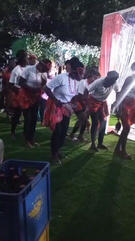 A group of women are dancing at an outdoor event on artificial turf. The women are barefoot and wear white shirts, black leggings, and patterned skirts. Each woman also wears a necklace and a headband. They are bent forward at the waist and moving together as part of their dance routine. Other people stand in the background, some watching the dancers. There are decorations including sheer draping, and green lighting on nearby trees. A blue crate full of beer bottles sits in the foreground.
