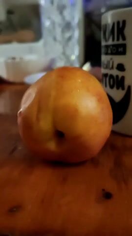 The video shows a close-up of a single nectarine on a wooden surface. The nectarine is round and has a reddish-orange skin with some yellow highlights. The surface appears to be a table. In the background, out of focus, there is a white mug with writing on it and what looks like a glass bottle or vase. The lighting is dim, creating a soft focus effect. The camera is stationary.