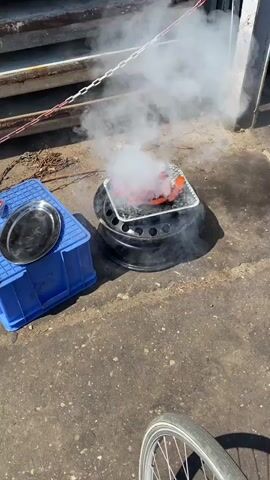 A video shows a close-up of hot dogs being grilled over an open fire. The grill is a makeshift setup, using a car tire as a stand for a metal tray filled with hot dogs. Smoke billows from the grill, obscuring the hot dogs at times. In the background, a blue plastic crate and a metal platter are visible. The video then pans out to show a large pile of old tires, some of which appear to be burning. A shopping cart is also visible, filled with debris. The scene appears to be an outdoor junkyard or recycling facility.