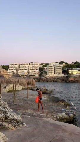 A young man wearing blue swim trunks and a dark top stands on a rocky shore, holding an orange flotation device. He is positioned near three thatched umbrellas and a pool ladder that descends into the dark water. In the background, several multi-story white buildings are visible, situated on a hill overlooking the water. The sky above is a soft gradient of pink and pale blue, suggesting either dawn or dusk.