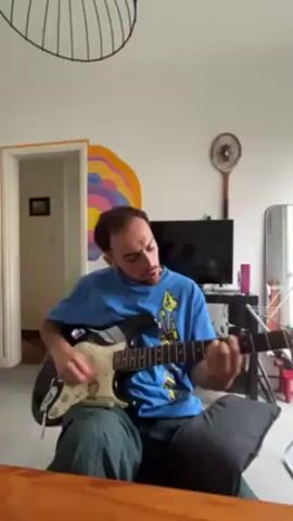 The video shows a man sitting on a couch and playing an electric guitar. He is wearing a blue t-shirt and green pants. The guitar is black and white. He appears to be singing while playing. Behind him is a TV, a mirror, and a painting on the wall. There is a doorway leading to another room in the background. A decorative lamp hangs from the ceiling.