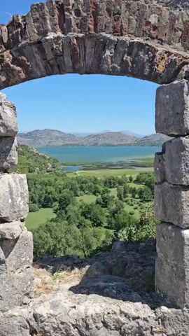 The video opens with a view framed by a stone archway, looking out onto a lush green landscape. In the foreground, weathered stones form the arch and the ledge below. Beyond the arch, a wide, calm body of water, appearing to be a lake, stretches into the distance, bordered by rolling hills and mountains under a clear blue sky. The landscape is dotted with trees and patches of green fields. The camera pans slowly to the right, revealing more of the expansive view, with the details of the distant hills becoming slightly clearer. The video appears to be a scenic landscape shot, focusing on the natural beauty of the environment. No people or specific actions are visible.