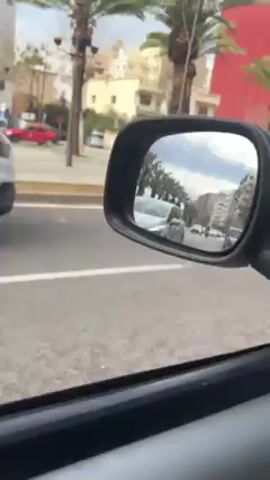 The video, shot from inside a car, captures the immediate surroundings as an ambulance passes.

At the beginning, a white ambulance with red and white flashing emergency lights on its roof is seen passing the car on the left side. The words "AMBULANCE" and "ASSISTANCE AMBULANCE" are visible on its side. In the background, palm trees line the street, and a reddish-orange building, along with other urban structures, can be seen under a partly cloudy sky. The distinct wailing sound of an ambulance siren is prominent throughout this initial segment.

As the video progresses, the ambulance moves further ahead, and its rear becomes more visible. A white symbol, possibly a crescent moon, is present on the back of the ambulance. The rearview mirror of the car provides a reflection of the street behind, showing other vehicles. The camera then slightly shifts to show more of the car's interior, including a dusty dashboard, a circular air vent, and a portion of the steering wheel.

The ambulance continues to move away in the distance, visible through the car's windshield, as other cars also populate the road. The siren sound gradually diminishes as the ambulance disappears from view. The scene depicts a typical daytime street environment in what appears to be an urban area with a warm climate, given the presence of palm trees.