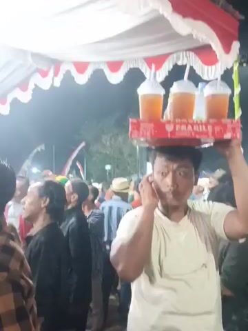 A young man is carrying a red tray laden with drinks on his head. The tray contains several plastic cups filled with a light brown liquid and straws, as well as several plastic bottles of water and other beverages. The man is wearing a light-colored t-shirt and is walking through a crowded outdoor area under a tent decorated with white and red fabric. People are visible in the background, some looking towards the man. The atmosphere appears to be that of an event or market.