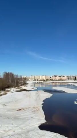 The video shows a landscape with a river flowing through a partially snow-covered area. The sky is clear and blue with a few faint clouds. In the background, there is a row of buildings, and further to the right, there are two large, dome-shaped structures with a metallic sheen. The river has open water areas amidst the melting snow and ice, and the water appears dark. On the riverbanks, there's a mix of snow and exposed ground, along with some trees. The camera angle suggests a slightly elevated position, offering a wide view of the scene.