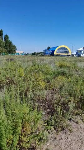 The video shows a long shot of a grassy field with a blue and yellow arched building in the background. The building appears to be a sports complex. There are trees and other smaller buildings visible to the left and right of the sports complex. The sky is clear and blue, indicating a sunny day. The camera is stationary and looking slightly upwards towards the building.