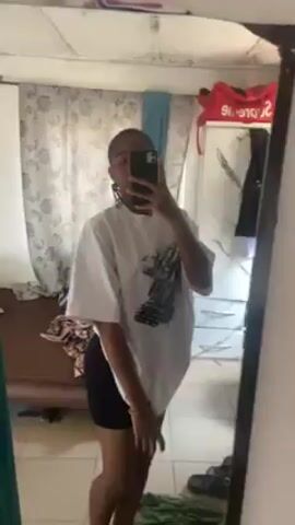 A young woman stands in front of a mirror taking a selfie. She is wearing a white oversized t-shirt with a graphic print and black shorts. Her hair is in braids. The room appears to be a bedroom, with a window covered by patterned curtains, a red Supreme bag on top of a piece of furniture, and a partially visible bed. The mirror has a black frame and is somewhat dirty. The woman holds her phone up to take the picture, obscuring her face. She slightly shifts her position while taking the selfie.