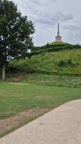 A video begins by showing a path leading through a grassy area. A large tree stands on the left with a bench in front of it. Further back, a grassy hill rises, crowned by a monument. The video then pans to the right, revealing more of the grassy park and a bandstand with red trim. Beyond the bandstand, trees and buildings are visible. The final shot shows a wide expanse of lawn with a few trees and distant buildings under a cloudy sky.