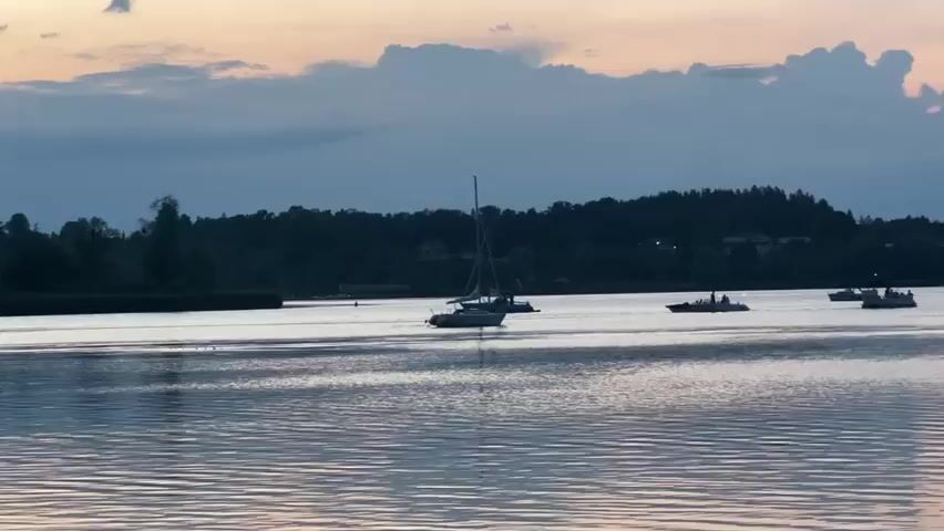 The video shows a body of water, likely a lake, with several boats on it. In the foreground, there is a wooden structure resembling a small house or boathouse with a deck. A person is standing on the deck, leaning over the railing and looking out at the water. Several boats, including sailboats and a speed boat with people on board, are visible on the water. The sky is overcast with clouds, and the lighting suggests it is either dawn or dusk. The overall scene depicts a tranquil evening or morning on the water.
