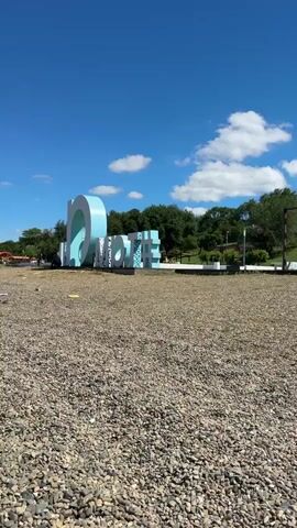 The video displays an outdoor scene under a bright blue sky with scattered white clouds. In the foreground, a wide area is covered with small, multi-colored pebbles or gravel. Dominating the middle ground is a large, light blue and white sculpture spelling out the word "LOVE," with individual letters clearly visible. This sculpture is situated on a paved surface.

Behind the sculpture and extending to the right, there is a green, tree-covered hillside. To the far right, a wooden pergola structure with several upright posts and crossbeams is visible. The camera performs a slow pan from left to right, offering a broader view of the setting. The scene appears to be a public area or park, likely a tourist spot, given the prominent "LOVE" sign.

The time of day is clearly daytime, indicated by the bright sunlight and the visibility of shadows. The weather is sunny and clear. No people are visible in the brief clip. The location context suggests this scene is in Taman, Russia.