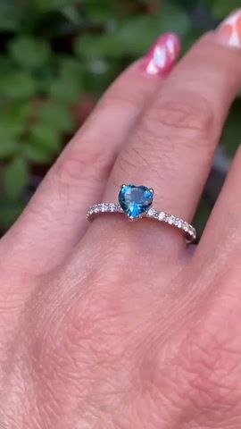 The video shows a close-up of a hand wearing a silver ring with a heart-shaped blue stone. The ring has small clear stones along the band. The background is blurred and shows green foliage. The hand appears to be moving slightly, causing the ring to catch the light. The fingernails are painted with colorful nail polish.