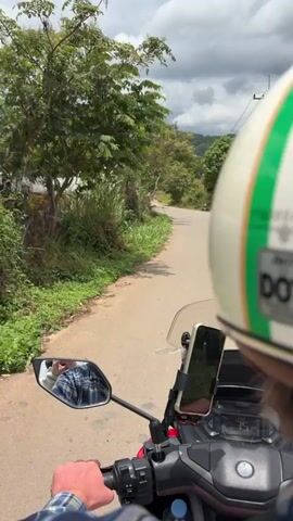 A first-person perspective video shows a person riding a motorcycle on a paved road. The rider's hands are on the handlebars, and a helmet is visible. A smartphone is mounted on the handlebars in front of the rider. The road is surrounded by lush green vegetation and trees. In the distance, there are hills and a cloudy sky. The rider is moving forward along the winding road.