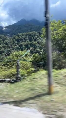 This video captures a scenic drive through a mountainous region, likely Bzypta, Abkhazia. The scene is dominated by lush green mountains covered in dense trees, with a cloudy sky overhead suggesting overcast or potentially rainy weather. In the foreground, a road is visible, indicating the perspective of a moving vehicle.

Several man-made structures dot the landscape. Early in the video, there are hints of a wire structure supported by stone pillars, possibly a trellis or an old fence. Further along, a tall metal power pylon stands prominently against the backdrop of the mountains. Adjacent to the pylon, there are modern-looking dark buildings, some appearing under construction with piles of gravel and building materials visible. A black car is parked near one of these buildings. A black metal fence with horizontal slats runs along the edge of one of the properties.

The overall impression is one of a rural or developing area nestled within a natural, mountainous environment. The lighting suggests it is daytime, but the cloud cover might be dimming the sunlight. There are no people clearly visible in the frame. The atmosphere is one of a journey through a landscape that blends natural beauty with emerging infrastructure.