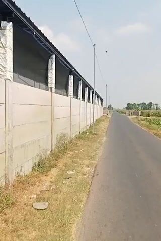 The video shows a long, narrow, paved road with dry grass and sparse vegetation on the left side. A concrete wall runs along the left side of the road, bordering a long, low-rise building with a dark roof and dark siding. The building has white, cylindrical elements hanging from its edge. The wall is made of large, light-colored concrete panels. On the right side of the road, the landscape is relatively flat with some vegetation and distant structures. A single utility pole with wires is visible along the road. In the distance, a motorcycle with a rider is approaching on the road. The sky is light blue with some clouds.