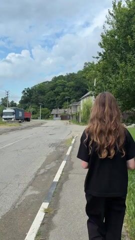 A person walks down a sidewalk next to a road. The person is seen from behind and wears a black t-shirt and black pants. The person has long, wavy, brown hair. The road has some potholes. A white truck with a blue shipping container and a red trailer is parked on the left side of the road. There are houses and trees in the background. The sky is cloudy.