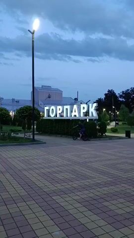 The video begins with a shot of a sign that reads "ГОРПАРК" in large, illuminated letters. A lamppost stands to the left of the sign, and a person on a bicycle is visible riding past in the foreground. The ground is paved with a pattern of reddish-brown and beige square tiles. The sky is overcast and appears to be dusk. The camera then pans to the right, showing a pathway with more people walking and a few bicycles parked. Trees line the path, and more lampposts are visible. The scene then shifts to a wider view of a park-like area with trees, flower beds, and people strolling. Further to the right, another large illuminated sign is visible, this one reading "МАЙКОП" with a red apple as the last letter. Several people are walking in front of this sign. The overall impression is an outdoor public space, likely a park, in the evening, with people enjoying the area.