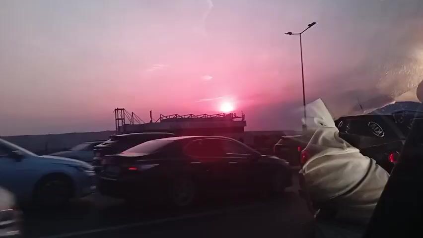 The video shows a sunset over a body of water, with a boat and a bridge in the background. The sky is a vibrant pink and orange, and the sun is setting behind the boat. There are cars on a road in the foreground, and the video appears to be taken from inside a car. A person's head is visible in the upper right corner of the frame.