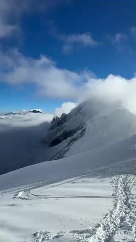 A panorama of a snowy mountain range unfolds under a bright blue sky dotted with white clouds. In the foreground, a vast expanse of snow is etched with tracks, suggesting recent activity. To the left, a low bank of clouds partially obscures the lower mountain slopes, revealing glimpses of rugged peaks and dark rock faces. Further into the scene, a cluster of tents, possibly a base camp, is visible on a snow-covered plateau. Several figures, dressed in outdoor gear, are present around the tents and scattered across the snow. In the distance, majestic, snow-capped mountains rise, their summits partially shrouded in mist, creating a dramatic and awe-inspiring landscape. The overall impression is one of a remote and challenging alpine environment.