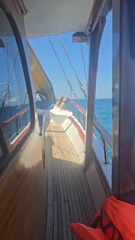 The video pans across the deck of a wooden boat on a sunny day. The sea is a vibrant blue with gentle waves. Part of a yellow slide is visible on the left side of the boat. The deck is made of wooden planks, and there are white benches with blue cushions. A wooden railing with metal supports runs along the side of the boat. The sky is clear and blue. The camera moves slowly, showing the different elements of the boat and its surroundings. There are no people visible in the video.