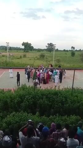 The video shows an outdoor gathering of people on a paved court, possibly a sports court, bordered by a fence and lush green bushes in the foreground. Several individuals, including men and women dressed in traditional attire, are seen conversing and interacting in small groups. Some individuals are standing near the center of the court, while others are positioned around the perimeter. In the background, there is a grassy field with scattered trees and a few light poles, suggesting a rural or semi-rural setting. A group of spectators is visible in the lower portion of the frame, seated and looking towards the court. The sky is overcast with clouds. The overall impression is of a community event or social gathering taking place outdoors.