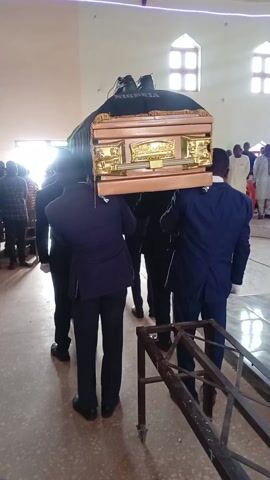 The video depicts a solemn procession inside a large hall or church. Several men, acting as pallbearers and dressed in dark suits, are seen carrying a light brown or golden wooden casket on their shoulders. The casket is adorned with intricate gold-colored decorative panels on its sides. A dark, possibly black, fabric is draped over the top of the casket, prominently displaying the word "NIGERIA" in white or light-colored letters. A microphone or recording device is also placed on top of the draped cloth.

Natural light floods the interior from large, high-set windows, suggesting it is daytime. The floor appears to be a light-colored, possibly tiled or polished surface. In the background, other individuals are visible, some standing and others seated, observing the procession. A metal trolley or stand is partially visible in the foreground to the right. The scene conveys the gravity and formality of a funeral or memorial service taking place in Ladura Foron, Nigeria.